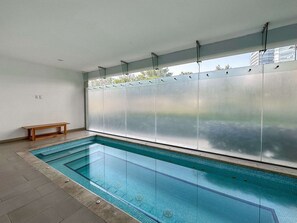 An indoor swimming pool with a bench, clear glass walls, and a view of buildings outside.