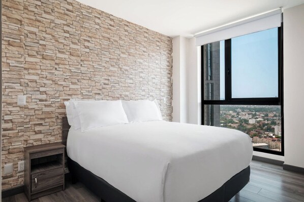 A modern bedroom with a stone wall, a large bed, a nightstand, and a city view through a large window.
