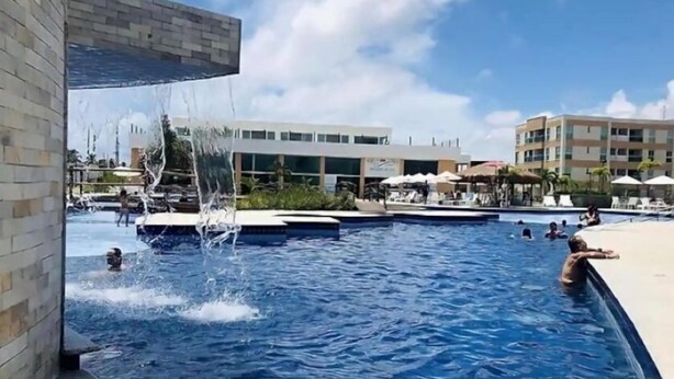 Una piscina con múltiples elementos acuáticos, como una cascada y chorros, rodeada de zonas de descanso y un edificio moderno al fondo.