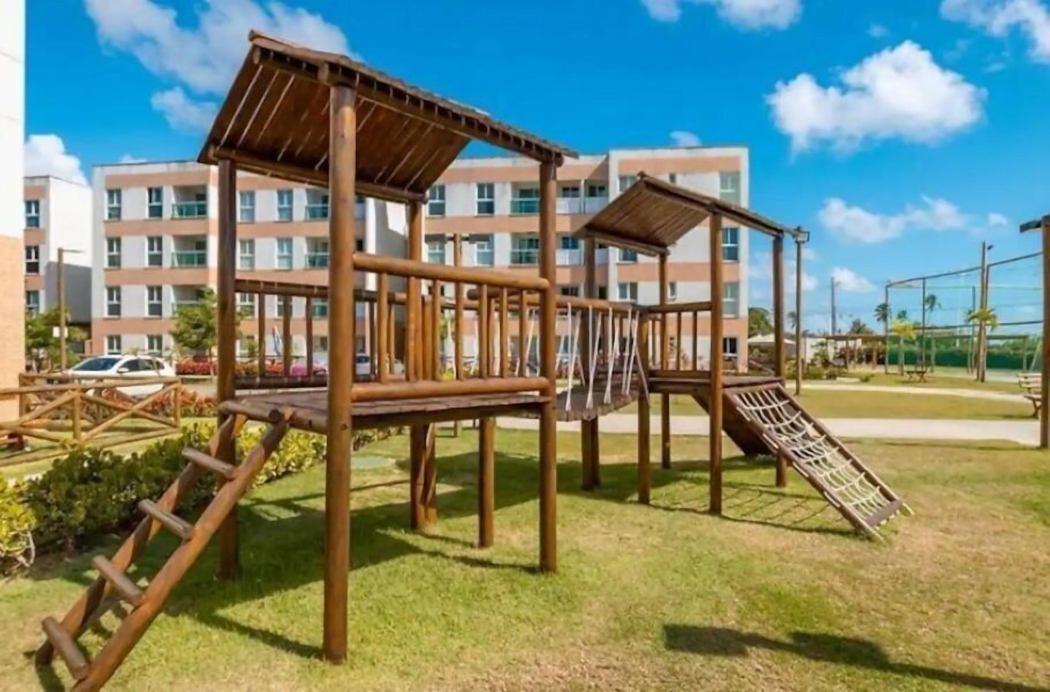 Un parque infantil con estructuras de madera y un tobogán, rodeado de césped y un edificio al fondo.