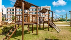 Un parque infantil con estructuras de madera y un tobogán, rodeado de césped y un edificio al fondo.