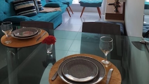 Una sala de estar moderna con un sofá, una mesa de comedor preparada para dos personas y un amplio ventanal con cortinas.