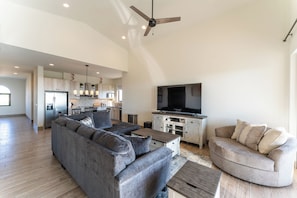 A spacious living area with a large gray sectional sofa, a smaller gray sofa, and a wooden coffee table. A flat-screen TV is mounted on a wooden cabinet, and a ceiling fan is visible.