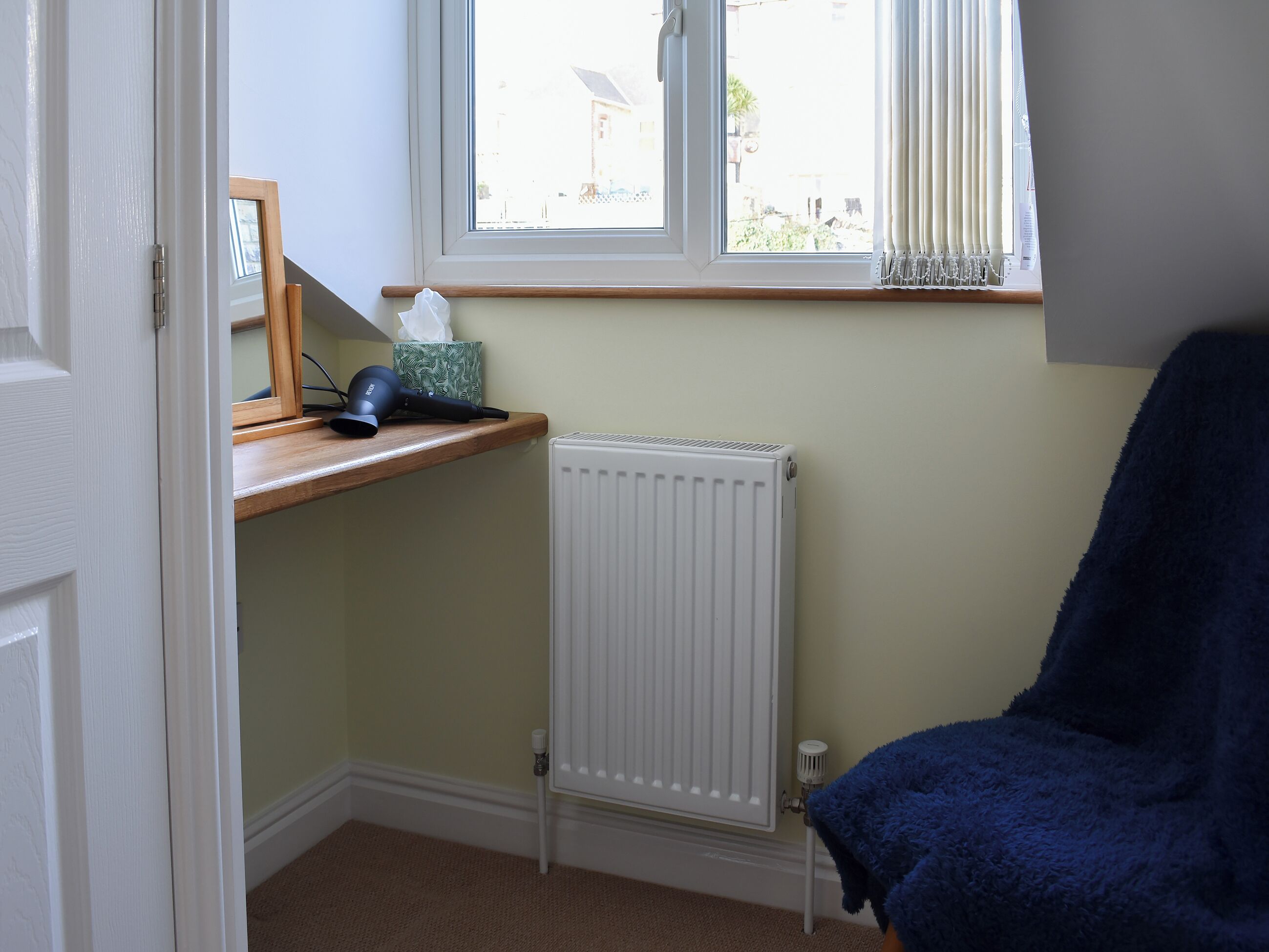A room with a radiator, a window with curtains, a shelf with a hairdryer, and a blue chair.