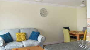 A living room with a grey sofa, yellow and blue cushions, a wooden coffee table, a dining table with chairs, and a wall clock. - 1 bedroom accommodation in Ventnor (Ventnor)
