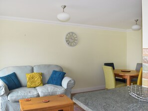 Una sala de estar con un sofá gris, cojines amarillos y azules, una mesa de centro de madera, una mesa de comedor con sillas y un reloj de pared.