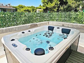 Un bain à remous doté d’un panneau de commande, entouré d’une bordure en béton, installé sur une terrasse en bois avec un jardin en arrière-plan.