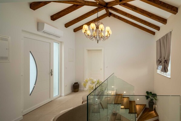 Un interior moderno con una escalera de cristal, vigas de madera, una araña y una puerta blanca.