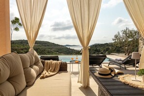 Un área de estar al aire libre iluminada por el sol, con piscina, vistas a colinas y una mesa con bebidas y un sombrero.