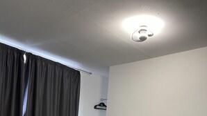 Un lit bien fait, avec une tête de lit blanche, une table de chevet sur laquelle se trouve une lampe, et un rideau noir partiellement tiré.