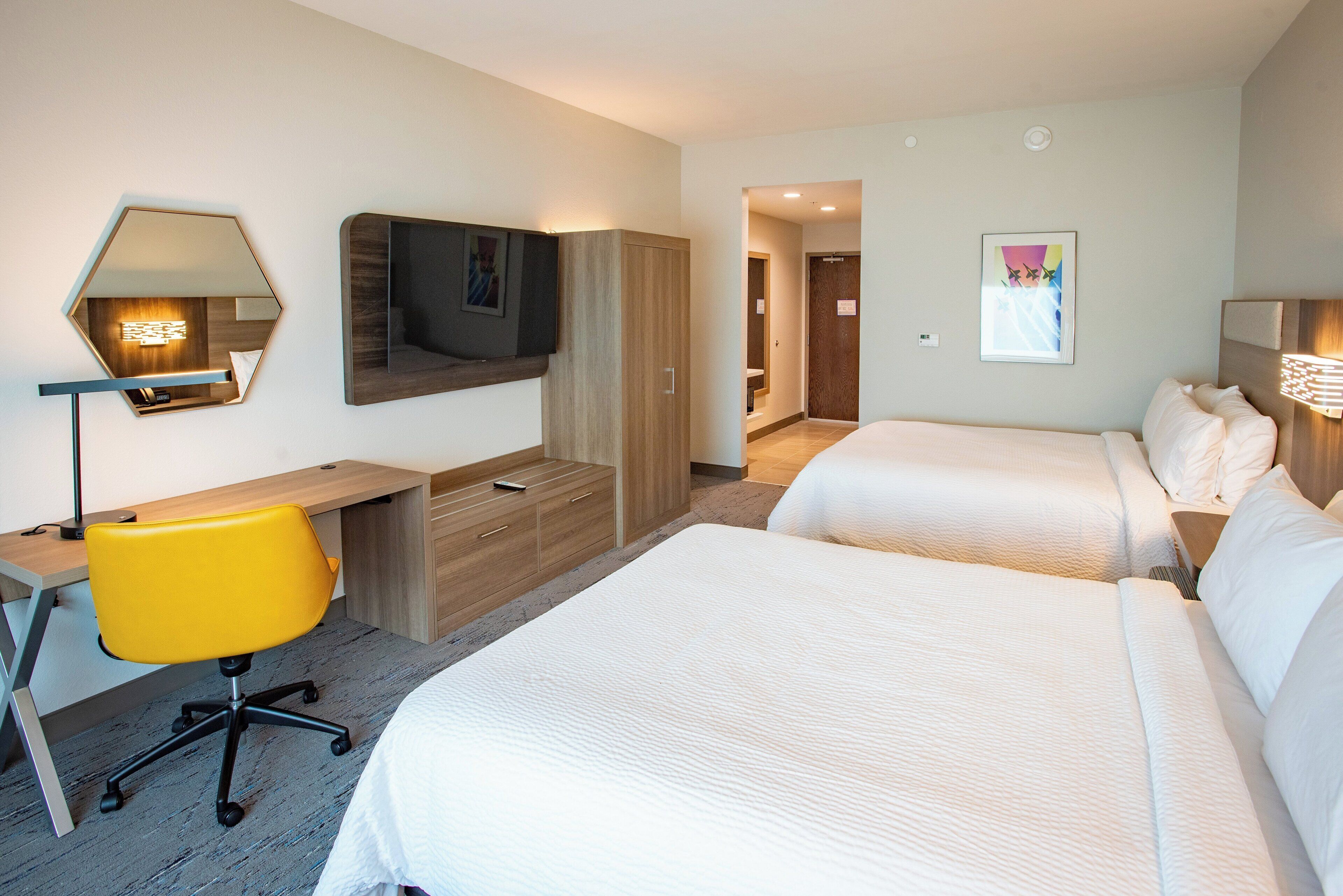 Una habitación de hotel con dos camas, un escritorio con silla, televisor y espejo.