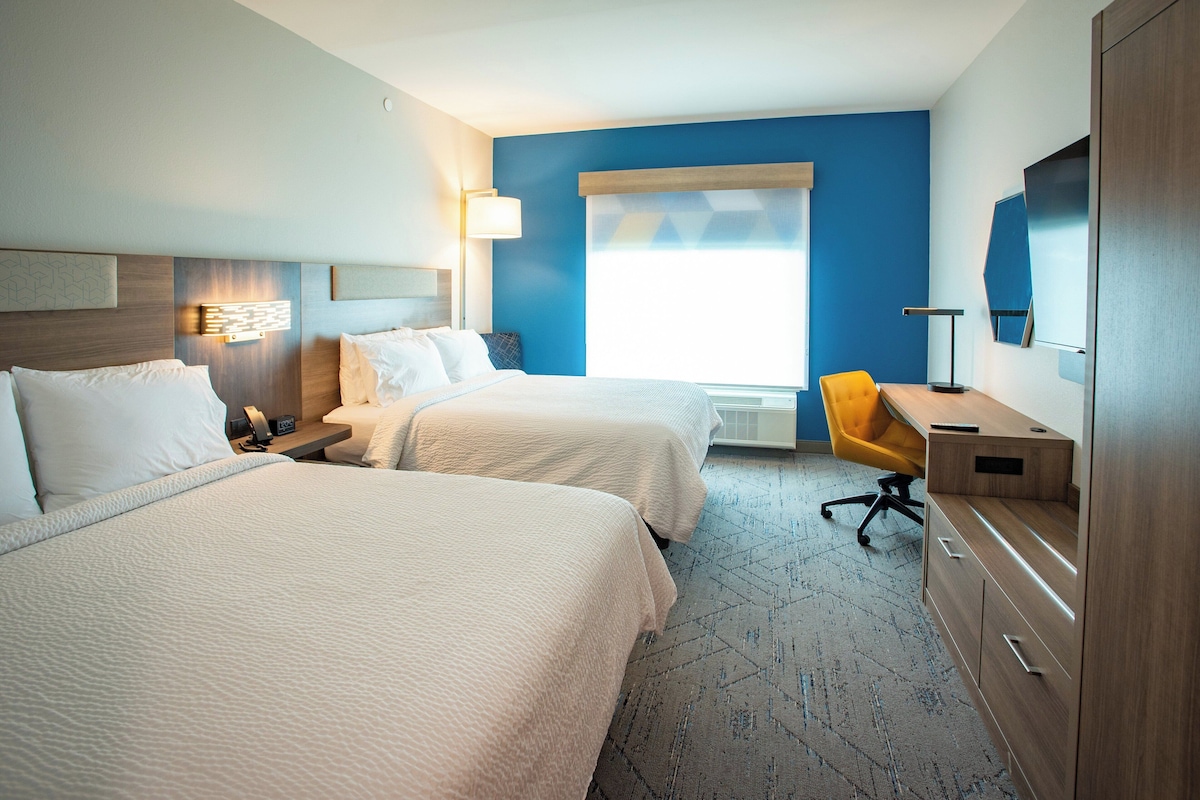 Une chambre d’hôtel avec deux lits, un bureau, une chaise, une télévision et une fenêtre avec des stores.