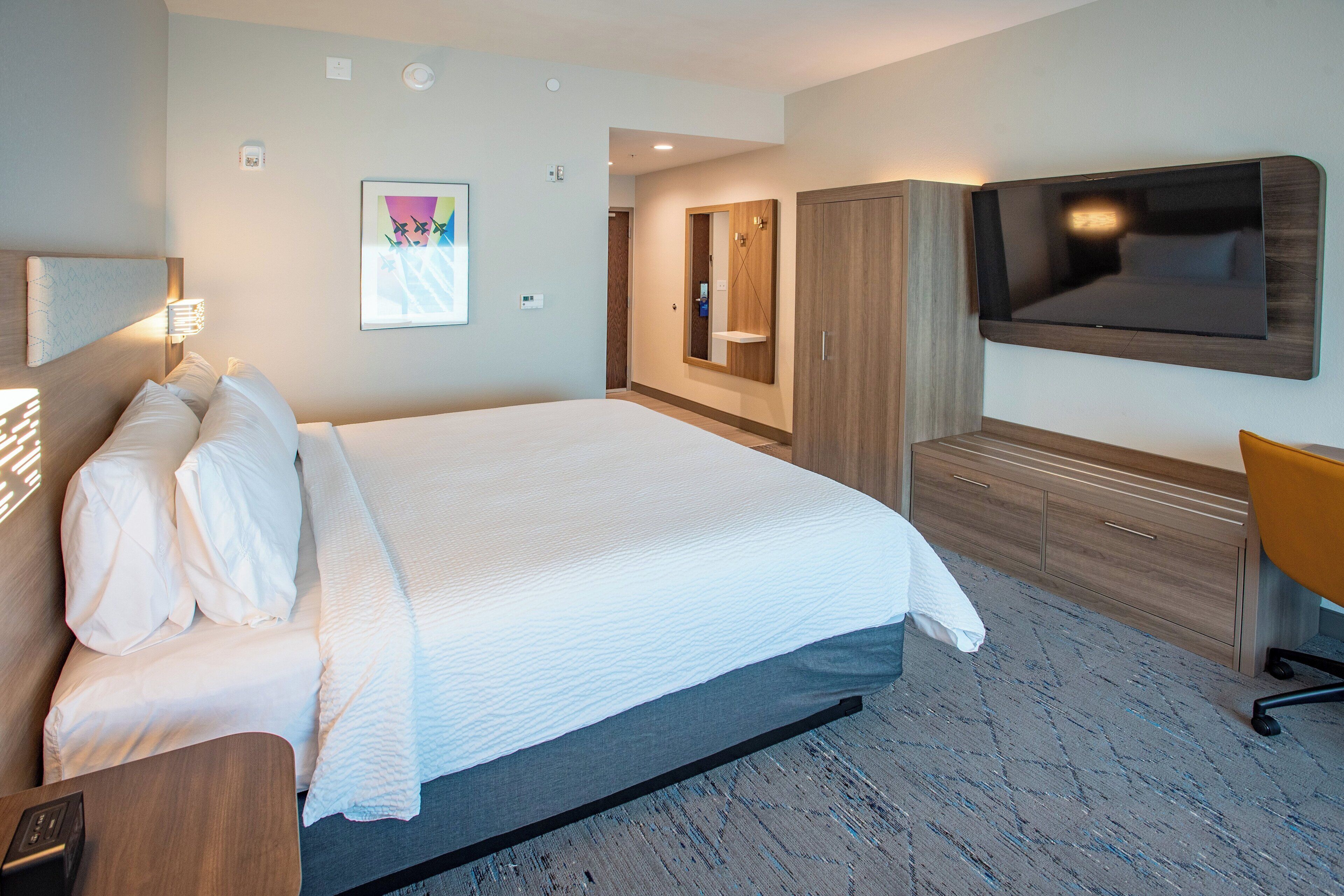 A hotel room with a large bed, a TV mounted on the wall, a wooden wardrobe, and a desk with a chair.