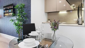 A modern dining area with a glass table, white plates, and clear glassware. A bamboo plant and a vase with flowers are on the table. - Charming 1-bed Apartment in London (Hayes)