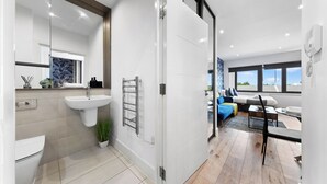 A modern bathroom with a toilet, sink, and towel rack. A glass door leads to a living area with a sofa, coffee table, and a view of the outdoors. - Charming 1-bed Apartment in London (Hayes)