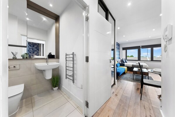 A modern bathroom with a toilet, sink, and towel rack. A glass door leads to a living area with a sofa, coffee table, and a view of the outdoors.