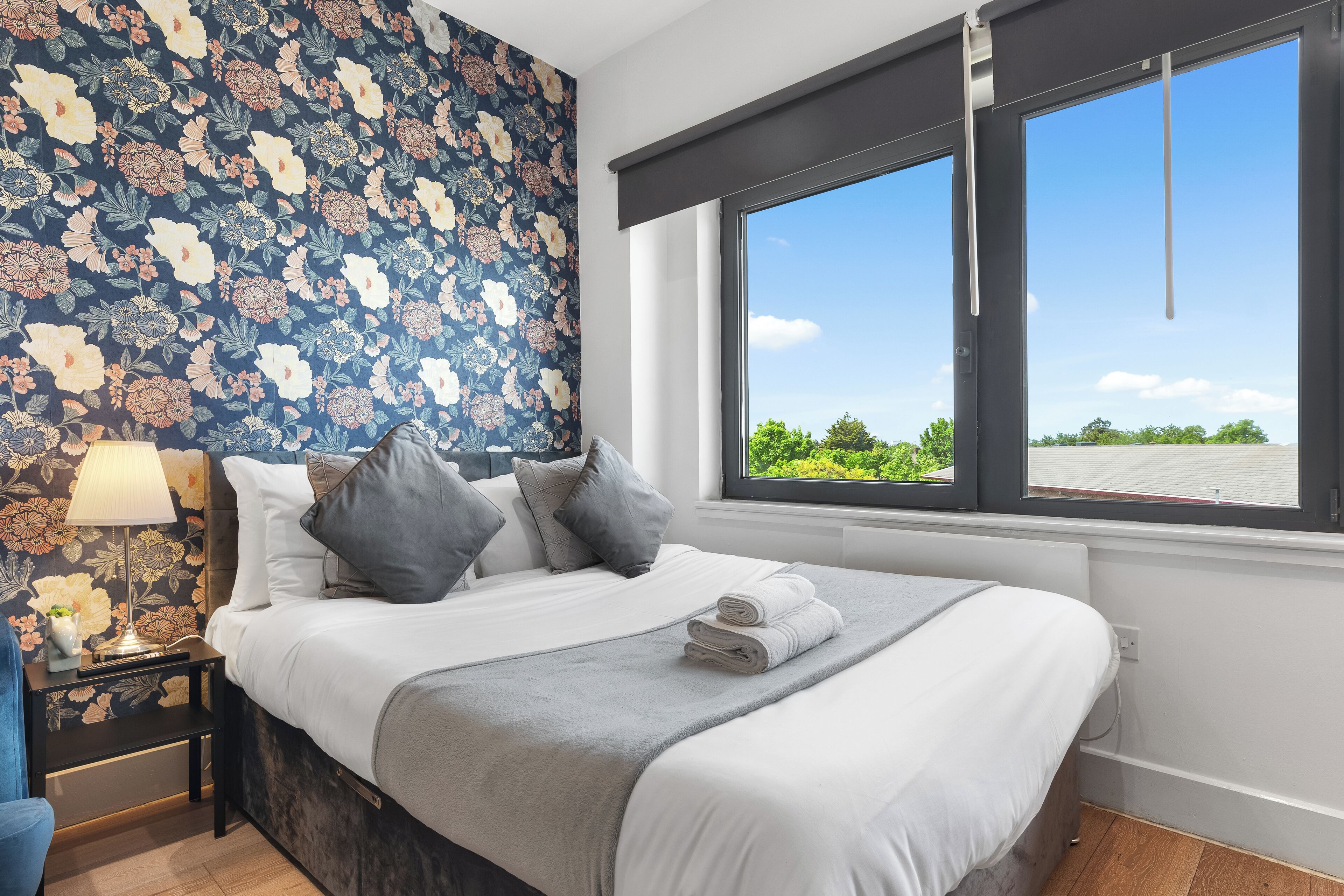 Un lit bien fait, recouvert d’une couverture grise, un papier peint à motifs floraux, une table de chevet avec une lampe et une fenêtre donnant sur un paysage verdoyant.