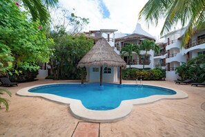 A swimming pool with a thatched roof structure, surrounded by lush greenery and a multi-story building with balconies.