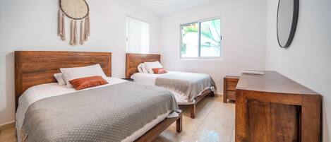 Un dormitorio con dos camas, un escritorio de madera, un espejo, un atrapasueños y una ventana con vistas a palmeras.