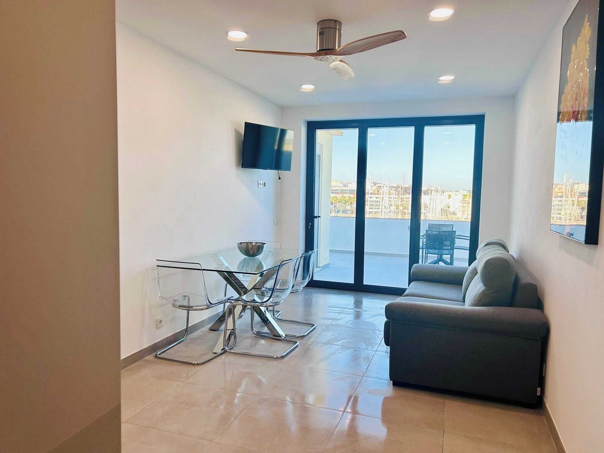 A modern living room with a glass dining table, a sofa, a ceiling fan, and a TV mounted on the wall.
