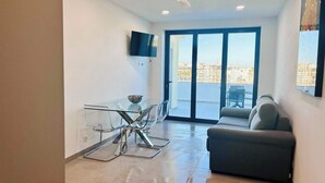 Una sala de estar moderna con una mesa de comedor de vidrio, un sofá, un ventilador de techo y un televisor instalado en la pared.