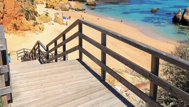 Una escalera de madera que conduce a una playa con agua azul cristalina y formaciones rocosas.