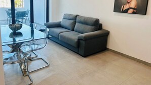 Un salon moderne avec un canapé gris, une table basse en verre et un coin repas comprenant une table et des chaises en verre. Une grande fenêtre offre une lumière naturelle et une vue sur l’extérieur.