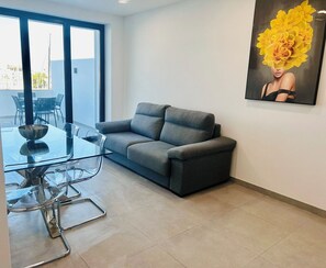 Uma sala de estar moderna com um sofá cinzento, uma mesa de centro de vidro e um espaço de jantar com mesa e cadeiras de vidro. Há uma janela grande que oferece luz natural e vista para o exterior.