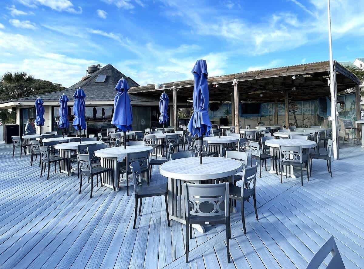 An outdoor dining area with round tables and chairs, covered by blue umbrellas, and a wooden deck.
