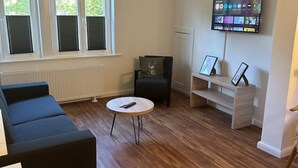A living room with a blue sofa, a round coffee table, a black armchair, a white bench with picture frames, and a mounted TV on the wall. - Apartment in Buchholz in der Nordheide, Germany (Buchholz in der Nordheide)