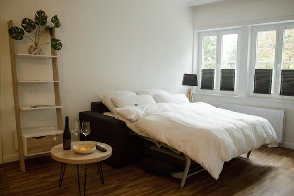 Une chambre à coucher avec un lit, une table de chevet sur laquelle se trouvent une bouteille et des verres, une petite plante et une fenêtre avec des volets.