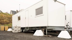Una casa móvil blanca con una ventana pequeña y una puerta, estacionada sobre un terreno de grava con una valla de madera y casas al fondo.