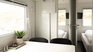 Un compartimento de tren moderno con interior blanco, una pequeña mesa con una planta, dos sillas negras, una cama con ropa de cama blanca y un televisor montado en la pared.