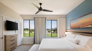 Ein Hotelzimmer mit einem großen Bett, einem Deckenventilator, einem Flachbildfernseher, einem Schreibtisch mit Lampe und einem Bild an der Wand.