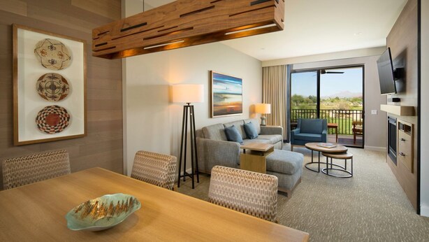 A modern hotel room with a dining area, a sofa set, a flat-screen TV, and a balcony with a view.