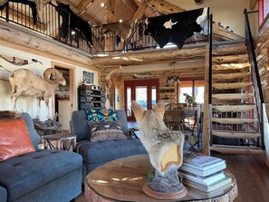 Een rustiek interieur met een vide, versierd met dierenhuiden, een zithoek met een bank en fauteuils, en een trap met houten leuning.