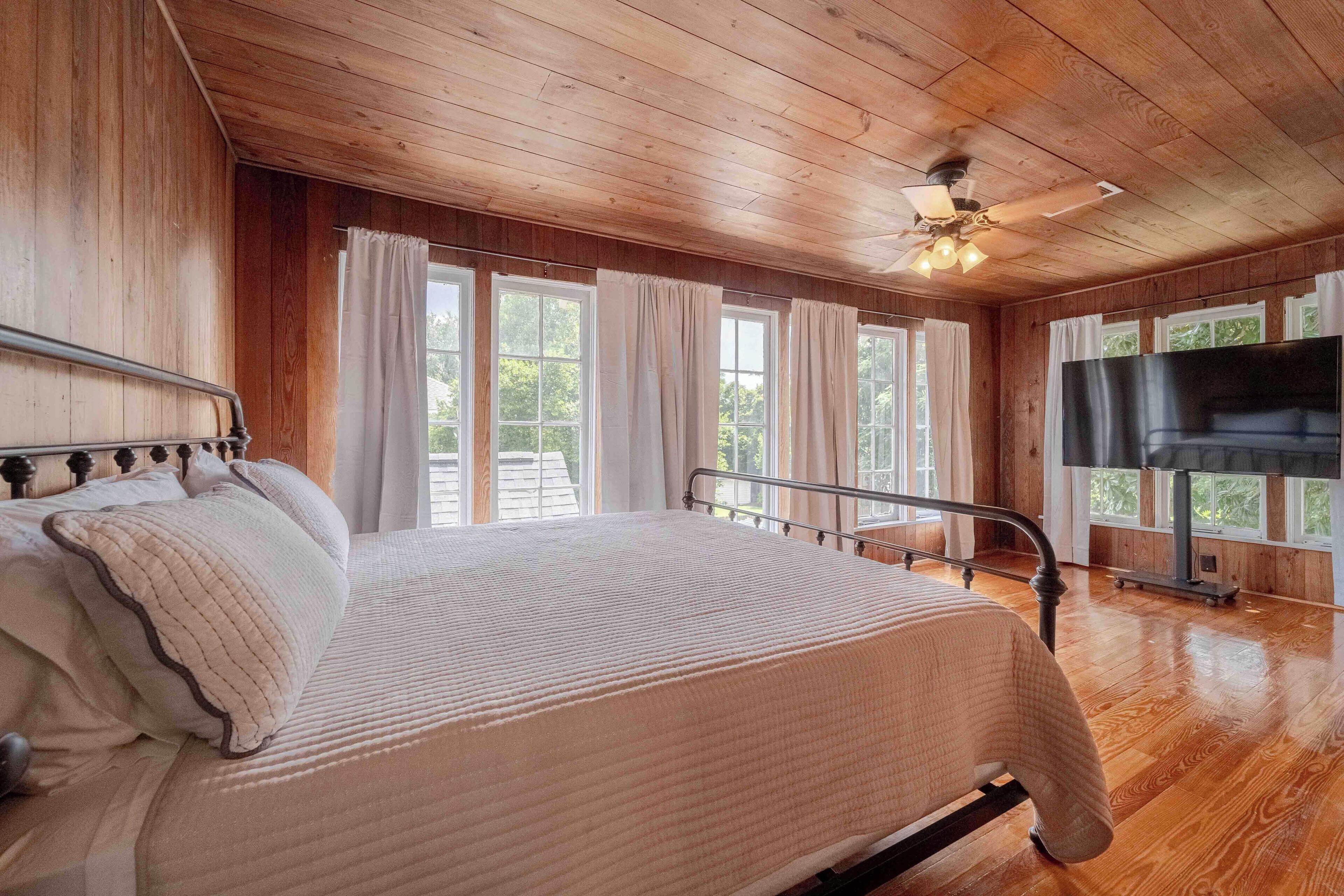 A wooden-paneled bedroom with a bed, curtains, a ceiling fan, and a TV.