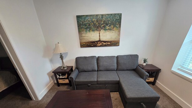 A living room with a gray sectional sofa, a dark wooden coffee table, and a painting of a tree on the wall. - Midtown Condo right on Main St! Walking distance to everything! (Lava Hot Springs)