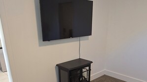 A room with a flat-screen TV mounted on the wall, a small black table with a lamp, and a dark wooden table. - Midtown Condo right on Main St! Walking distance to everything! (Lava Hot Springs)