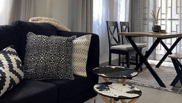 Una sala de estar moderna con un sofá negro, cojines estampados, una mesa de centro de madera y un comedor con mesa y sillas.
