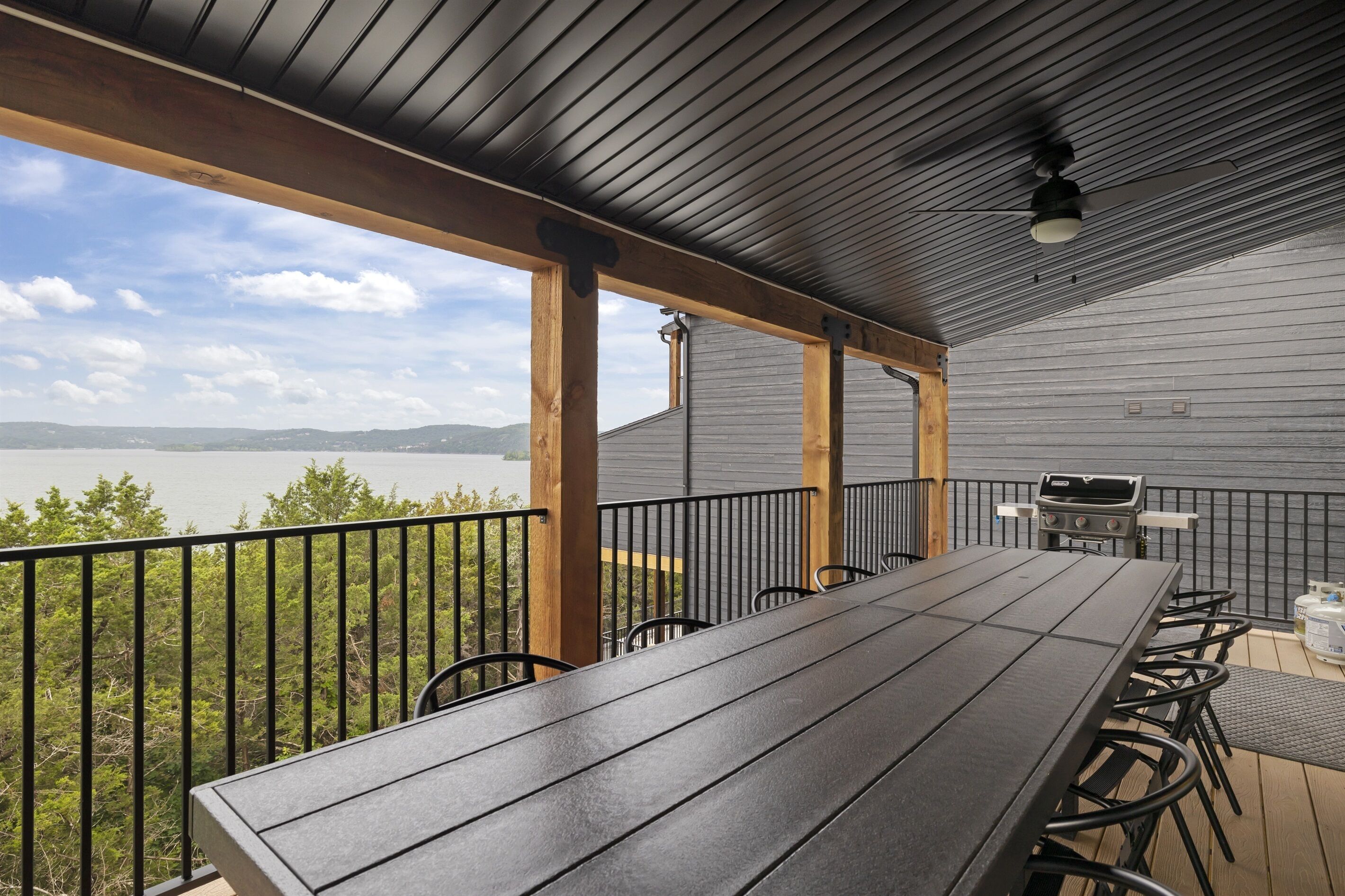Una terraza techada al aire libre con una mesa larga y sillas, una parrilla y vistas a un lago y montañas.