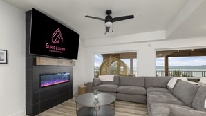 Moderni olohuone, jossa on takka, suuri harmaa kulmasohva ja litteänäyttöinen televisio, jolta näkyy ohjelma nimeltä ”Super Luxury Home Rentals”.
