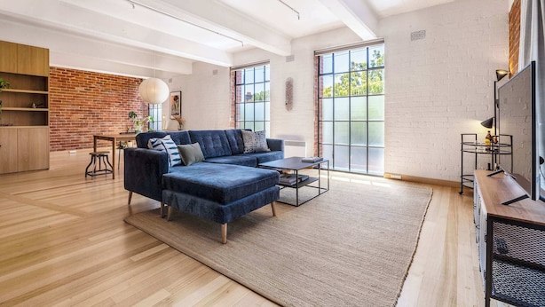 A spacious living area with a blue sofa, a coffee table, and a rug. There is a brick wall and large windows.