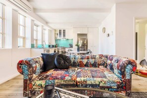 A living room with a colorful quilted sofa, a dining area with a table and chairs, and a kitchen with a teal backsplash.