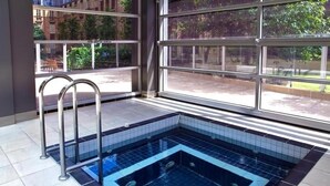 A square hot tub with a tiled floor, metal ladder, and large windows with a view of greenery outside. - Vintage glamour with indoor pool in heritage CBD (Melbourne)
