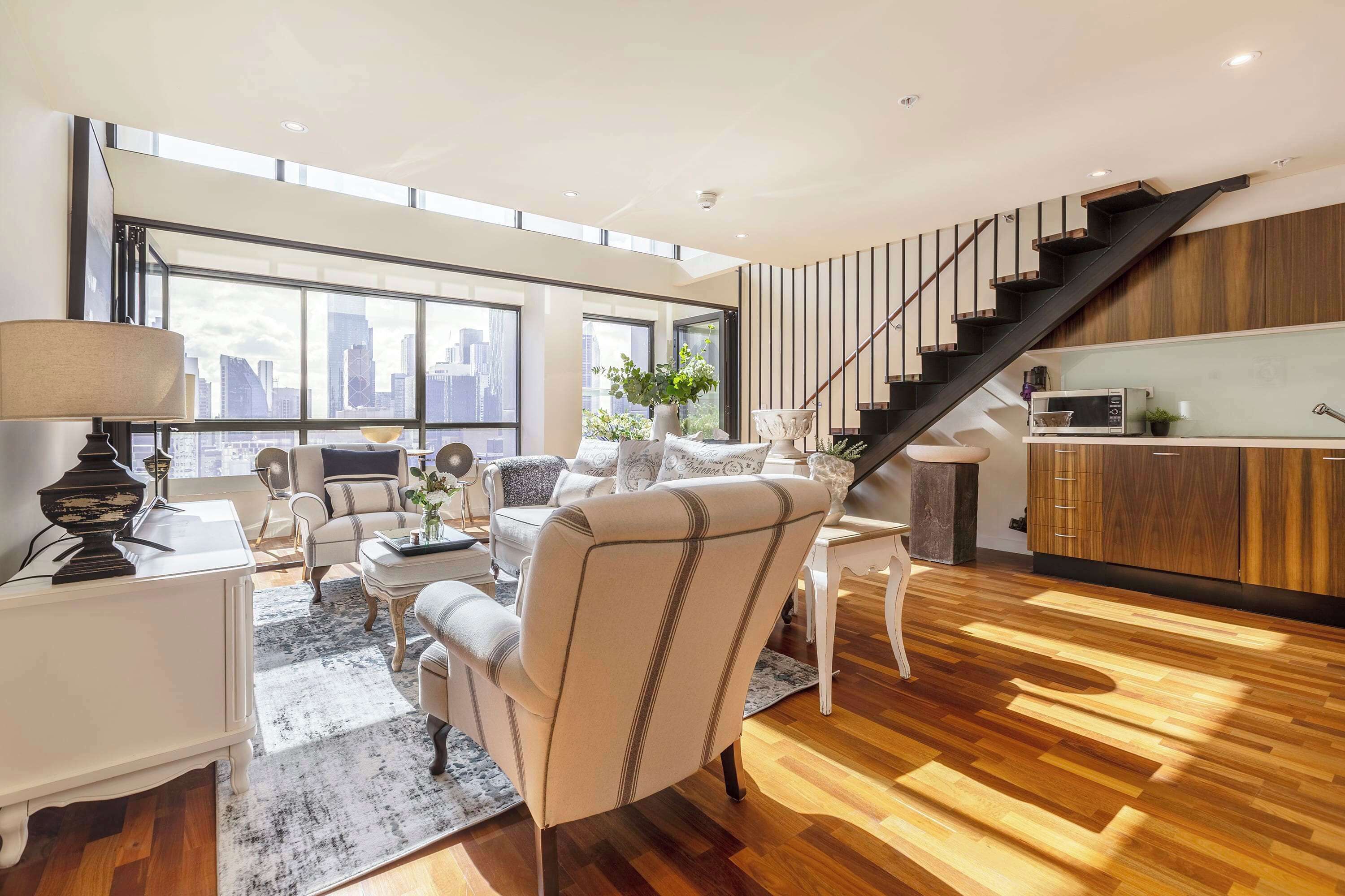A modern living room with a large window, a staircase, and a kitchen area.