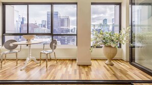 A room with large windows, a round table, two chairs, a potted plant, and a modern city view.