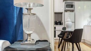 A modern room with a dark blue upholstered headboard, a crystal lamp on a dark dresser, a dining area with a white vase, and a kitchen with a microwave and refrigerator.