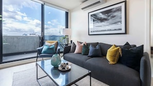 Ein modernes Wohnzimmer mit großem Fenster, einer Couch mit vielen Kissen, einem Couchtisch mit Dekoration und einem gerahmten Bild an der Wand.