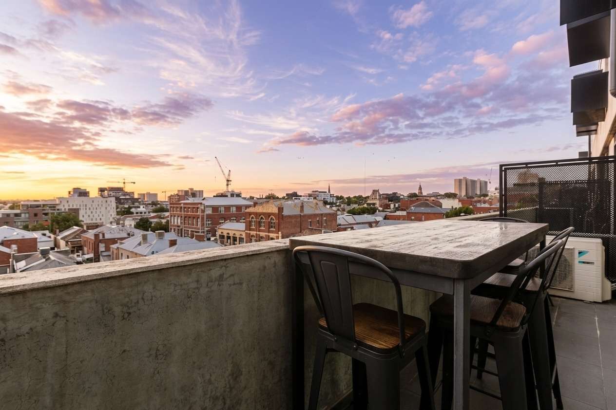 A rooftop terrace with a table and chairs, overlooking a cityscape with buildings and a clear sky.